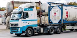 Halléns lastbil transporterar en tankcontainer från TAL längs en väg vid en industriell anläggning.