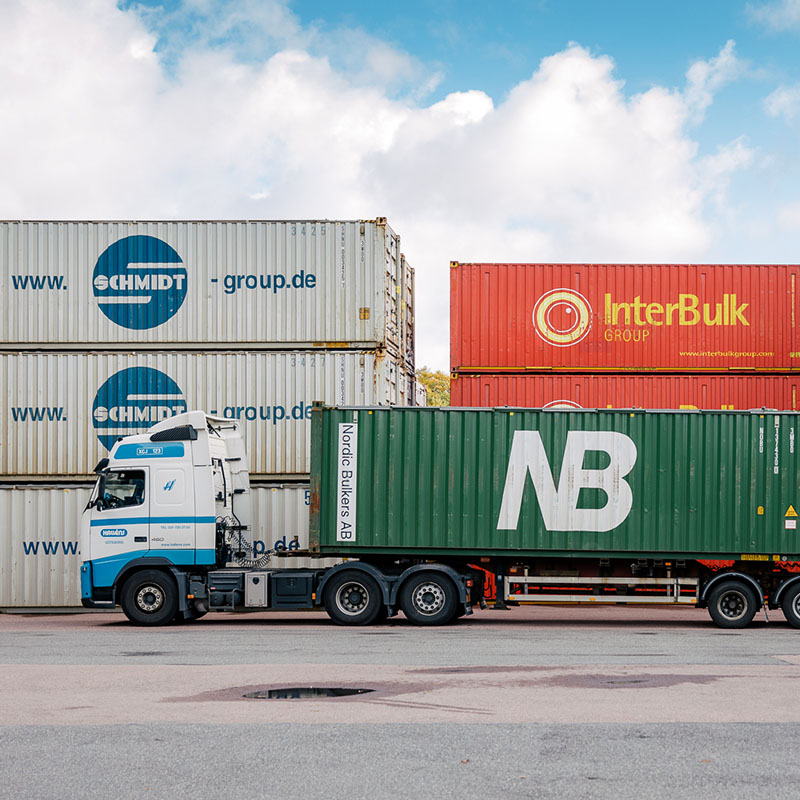 Halléns lastbil transporterar en grön NB-container framför staplade containrar från Schmidt och InterBulk Group