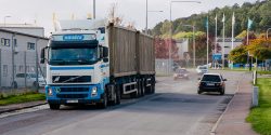 Halléns lastbil med en lång släpvagn kör på en industriell väg omgiven av grönska och byggnader.