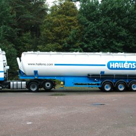 Halléns lastbil med en vit och blå tanktrailer parkerad framför en skog.