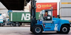 Blå Kalmar containertruck flyttar en grå container på en containerterminal, med fler containrar och en Halléns lastbil i bakgrunden.