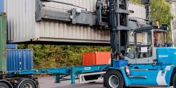 Blå containertruck lyfter en stor grå container och placerar den på en lastbilstrailer.